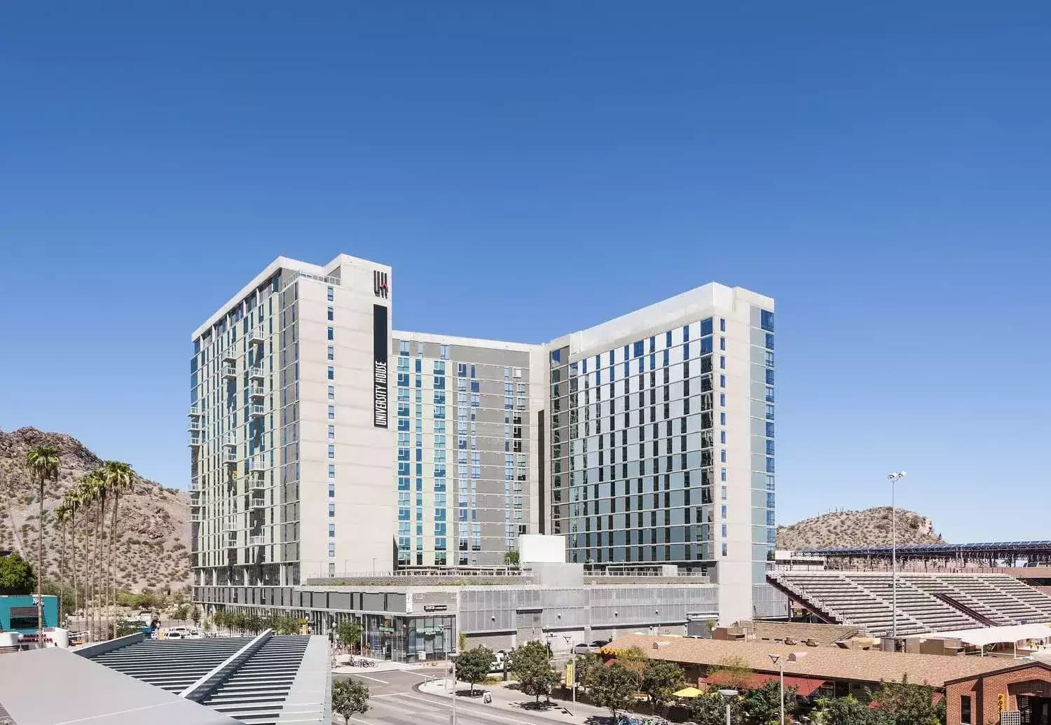 A modern high-rise hotel with glass windows stands under a clear blue sky near University House Tempe, with mountains and palm trees in the background and a stadium with bleachers to the right.