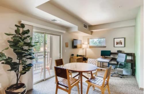 A bright living space at Flats at the Oval Fort Collins features a round dining table with four chairs, a potted plant by sliding glass doors, and a cozy work area with desk, chair, and computer in the corner by the window.