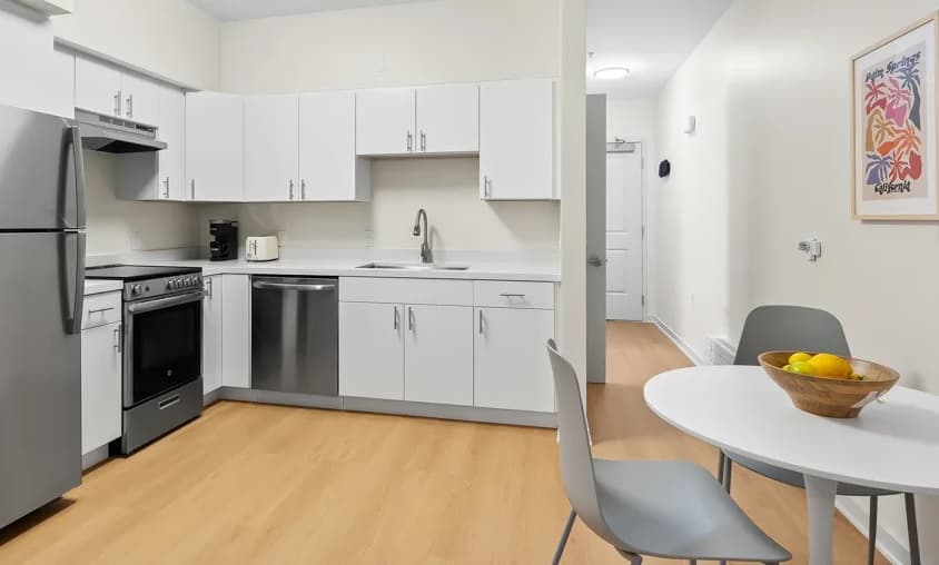 Modern kitchen at FOUND Study, featuring white cabinets, stainless steel appliances, and light wood flooring. A small round dining table with two gray chairs and a fruit bowl sits below a colorful framed picture—right in downtown Berkeley.