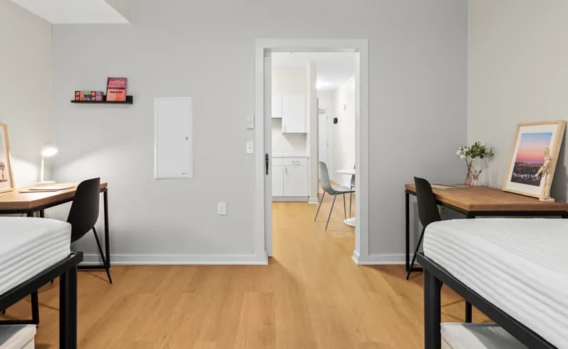 A bright, tidy dorm room at FOUND Study Downtown Berkeley features two beds, desks with chairs, and decor. The door opens to a kitchen area with white cabinets, a table, and two chairs on a wood floor.