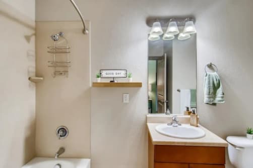 A modern bathroom at Flats at the Oval Fort Collins features a bathtub and shower, single sink vanity, wall mirror, shelf with a "Good Day" sign, and a neatly folded green towel on a ring. Four lights shine above the mirror.