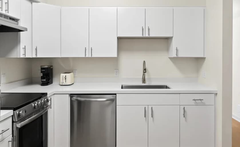 Modern kitchen at FOUND Study Downtown Berkeley features white cabinets, a stainless steel dishwasher, oven, and sink. A black coffee maker and white toaster sit on the countertop. The space is clean and minimalistic.