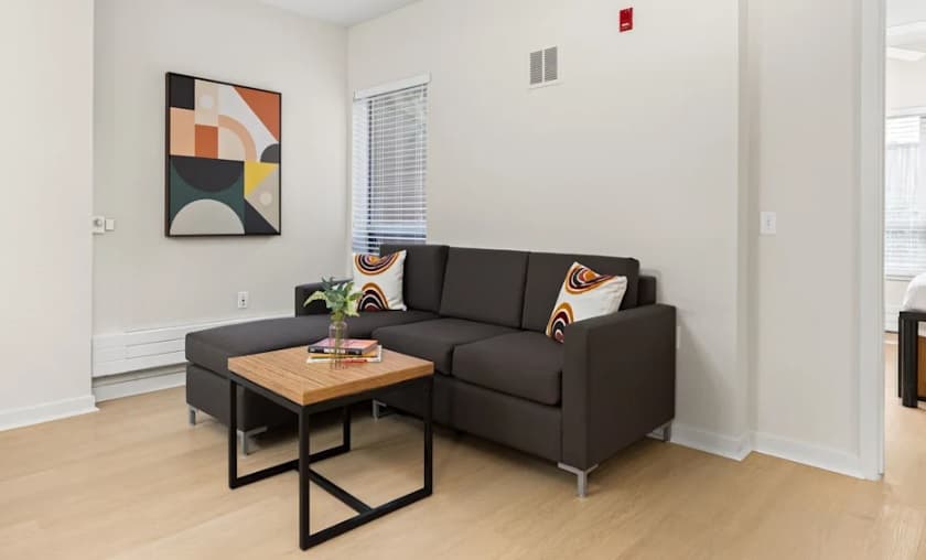 A modern living room at FOUND Study Downtown Berkeley features a dark gray sectional sofa, two decorative pillows, a wooden coffee table with a plant, abstract wall art, and light wood flooring. White walls and windows add brightness to the space.