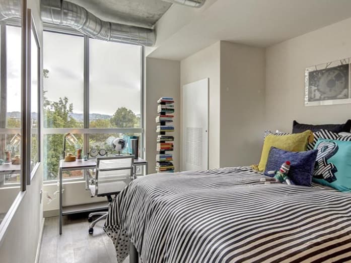Modern bedroom at 959 Franklin with large windows, a striped bedspread, colorful pillows, a small desk and chair, stacks of books, and urban views. Exposed ductwork and minimal decor enhance the contemporary vibe.