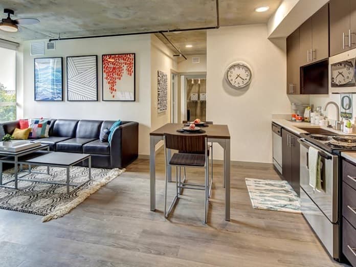 Modern open-concept apartment at 959 Franklin with a kitchen and living area. Features a black sofa, abstract wall art, clock, dining table with two chairs, and stainless steel appliances in the kitchen.