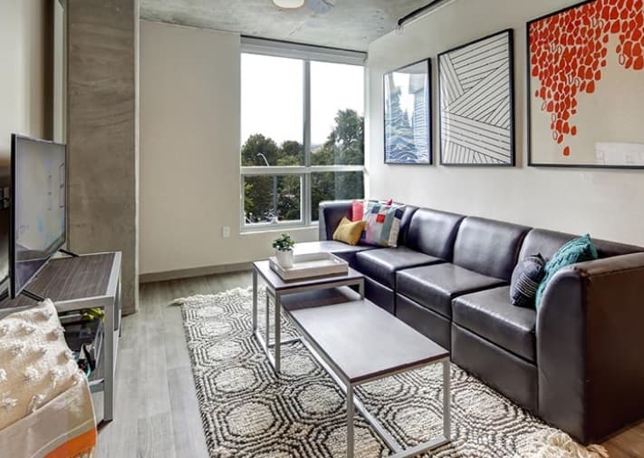 Modern living room at 959 Franklin with a black sectional sofa, geometric rug, two nesting coffee tables, and colorful wall art. A large window lets in natural light, and a TV sits on a stand opposite the sofa.
