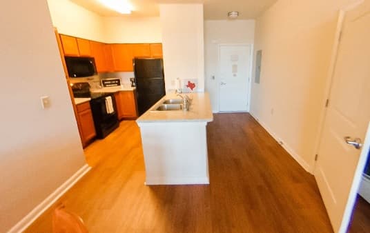 A modern kitchen in Gardens Apartments features wood cabinets, black appliances, and a central island with a sink. The space opens to a hallway with wood flooring and a white door at the end, perfect for stylish apartment rental living.