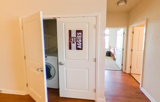 A partially open door reveals a laundry area with a washing machine. The door has a Texas A&M Aggies sign. Nearby, a hallway in The Gardens Apartments leads to well-lit rooms with wooden floors.
