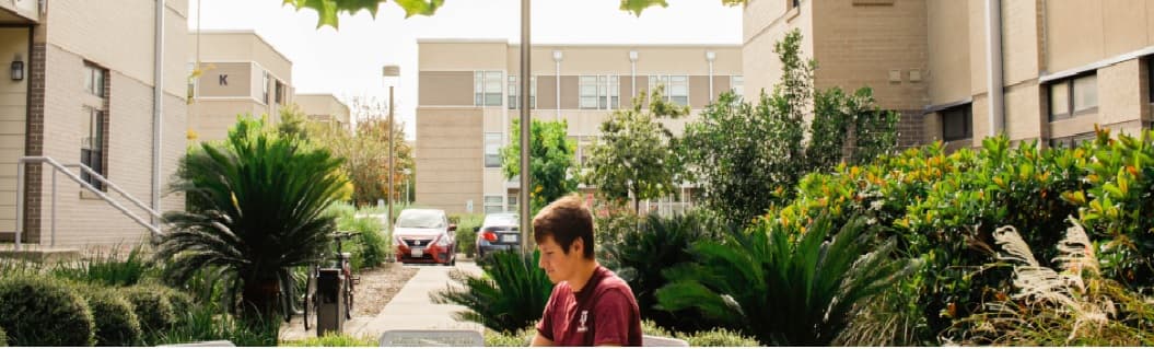 A young person sits at an outdoor table at The Gardens Apartments, surrounded by greenery and buildings, reading or working. Cars are parked in the distance along a pathway lined with plants and small trees.