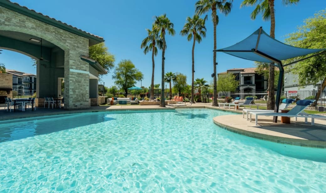 Outdoor swimming pool with clear blue water at Gateway at Tempe, surrounded by lounge chairs, palm trees, and shaded seating areas on a sunny day. Apartment buildings can be seen in the background.