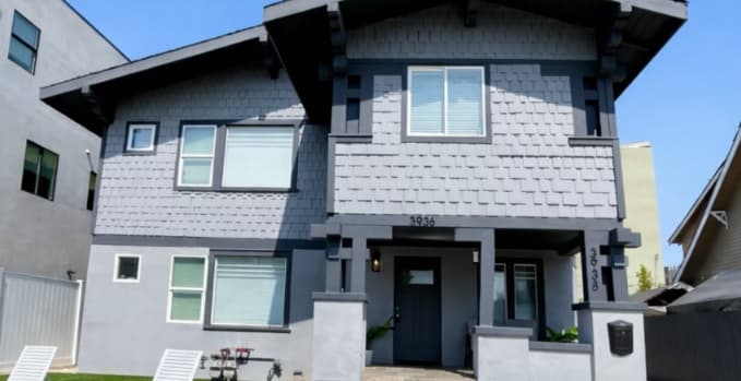 A two-story gray craftsman-style house with dark trim, a small front porch, and white-framed windows sits in the Galaxy Housing community. The front yard has green grass and a couple of white lawn chairs.