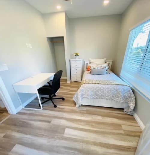 A small, bright bedroom in Mosaic Student Communities with a single bed, white desk and black chair, white chest of drawers, and window blinds. The light wood floor and simple decor make it ideal for student housing.