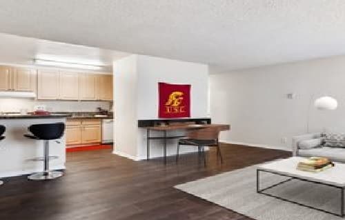 A modern student apartment with dark wood floors, a kitchen with light cabinets, a breakfast bar with two black stools, a desk adorned with a red and gold banner, and a gray sofa with a coffee table in the living area—ideal student housing.