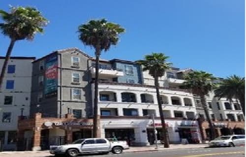 A street view of a multi-story building featuring student apartments on the upper levels, shops on the ground floor, palm trees lining the sidewalk, parked vehicles, and a clear blue sky above.