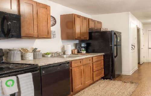 A modern kitchen at The Grove Apartments in Pullman features wooden cabinets, granite countertops, a black refrigerator, microwave, dishwasher, and neatly arranged kitchen items. The wooden floor is complemented by a rug in front of the sink.