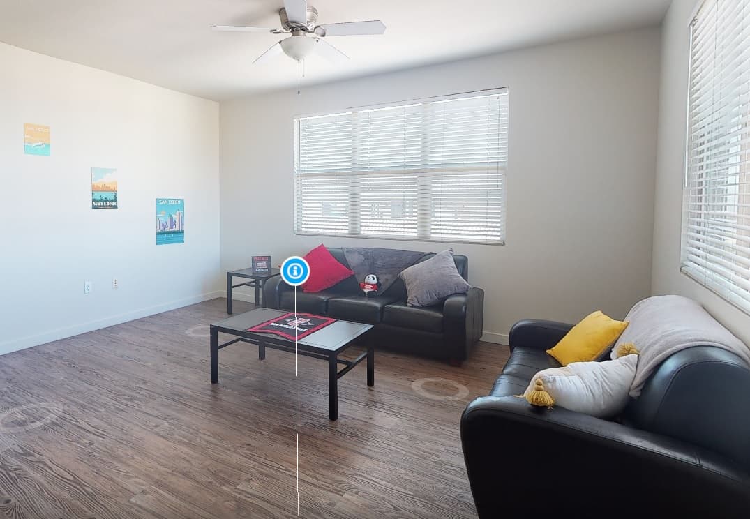 A bright living room at Granada on Hardy with two black sofas, a coffee table with magazines, and a side table. Large windows let in natural light, while throw pillows and colorful posters add charm near SDSU.