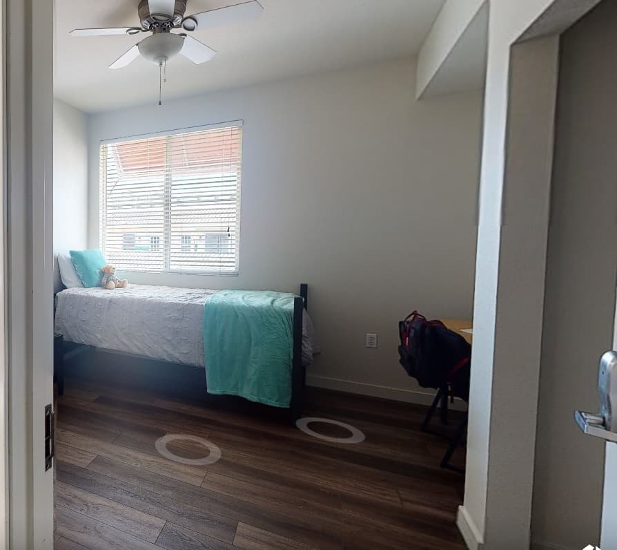 A small, tidy bedroom at Granada on Hardy with a single bed, white bedding, and teal accents. There’s a window with blinds, a ceiling fan, wooden floors, and a desk with a chair—perfect for SDSU students.