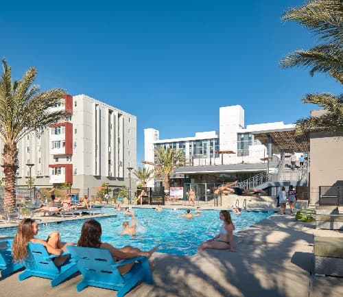 People relax and swim in a sunlit outdoor pool at Granada on Hardy near SDSU, surrounded by palm trees and modern apartment buildings, while others lounge on blue chairs under a clear blue sky.