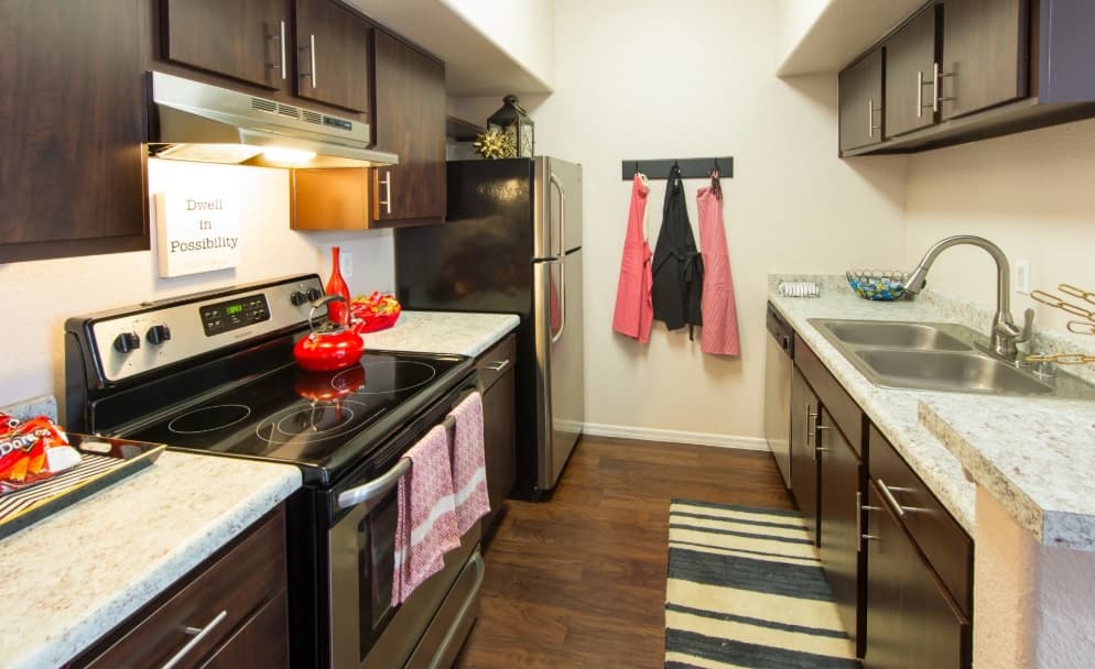 A modern kitchen at Gateway at Tempe features dark wood cabinets, stainless steel appliances, a double sink, and white countertops. Red accents, dish towels, and a striped rug add color, while two aprons hang near the refrigerator.