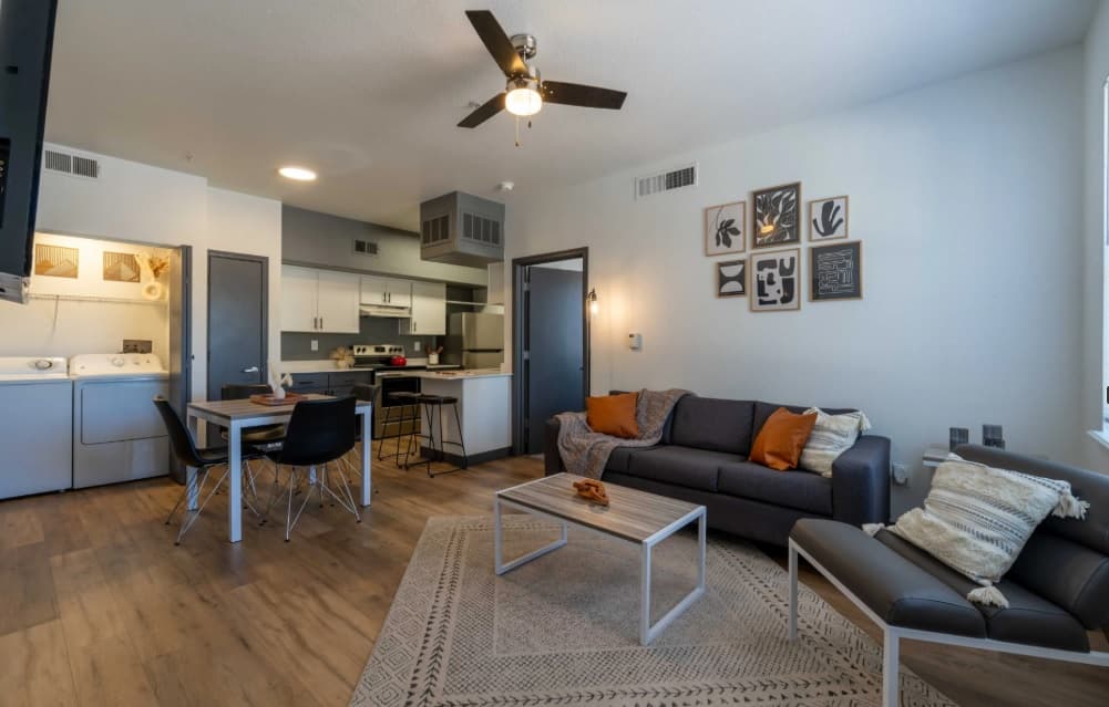 Modern apartment living room in Chico Student Housing with a gray sofa, armchair, coffee table, dining area, and open kitchen. Laundry machines in the corner, light wood flooring, neutral décor, wall art, and throw pillows complete the look.