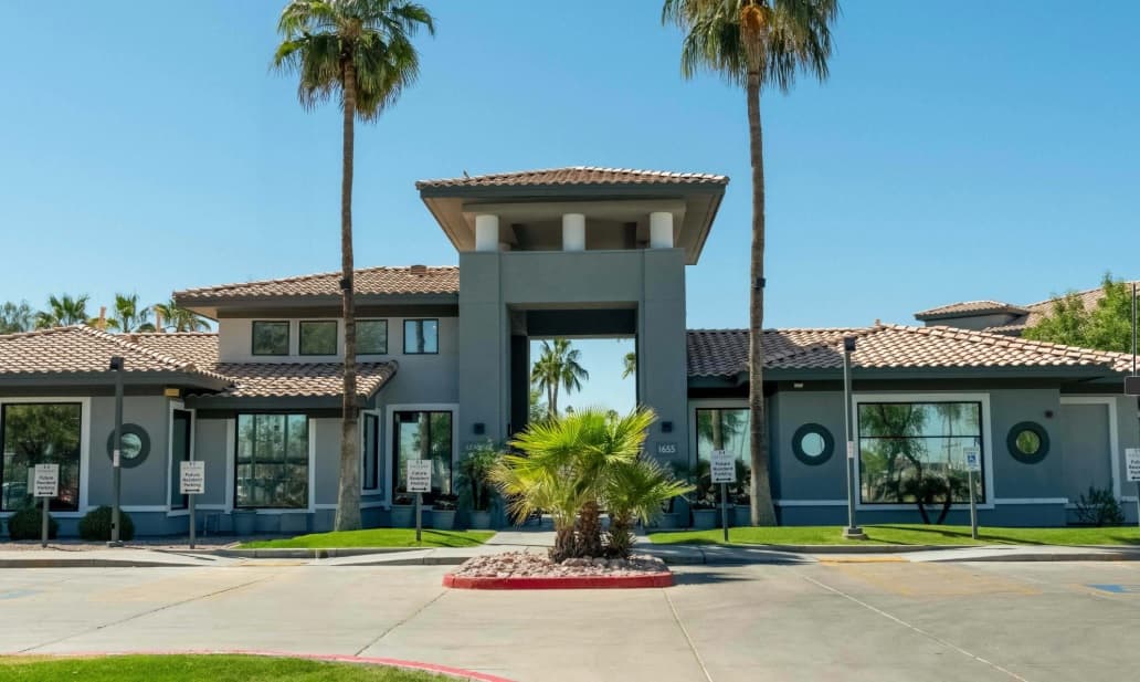 A modern building with a tiled roof, large central archway, and tall columns stands at Gateway at Tempe; gray exterior, large windows, palm trees, and manicured landscaping shine under a clear blue sky.