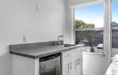 A small, modern kitchenette with white cabinets, a Heritage 2831 stainless steel countertop, a sink, and a mini fridge below; a window reveals a view of parked cars and buildings outside.