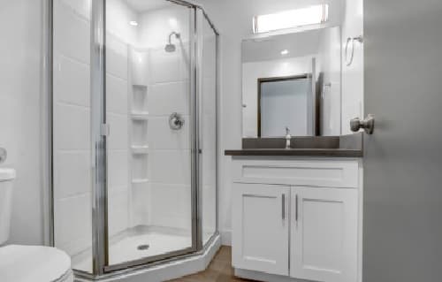 Modern bathroom with a glass-enclosed corner shower, white vanity with gray countertop and sink, wall-mounted mirror, and white walls. A Heritage 2831 toilet is visible in the left foreground.
