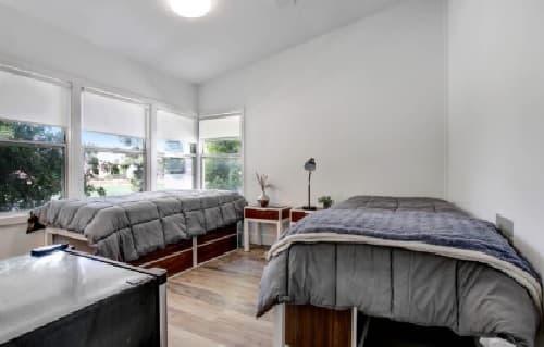 A bright Heritage 2831 bedroom with two single beds covered in gray bedding, a small desk with a lamp and plant between them, large windows with white shades, and wood flooring.