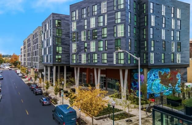 A modern mid-rise apartment building, HERE Seattle features large windows and a colorful mural at street level, overlooking a tree-lined urban street with parked cars.