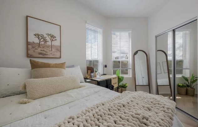 A cozy bedroom with a neatly made bed, beige and cream bedding, framed desert landscape art, a desk by two windows, a full-length mirror, and a mirrored closet. Plants and natural light add warmth in this inviting HERE Reno space.