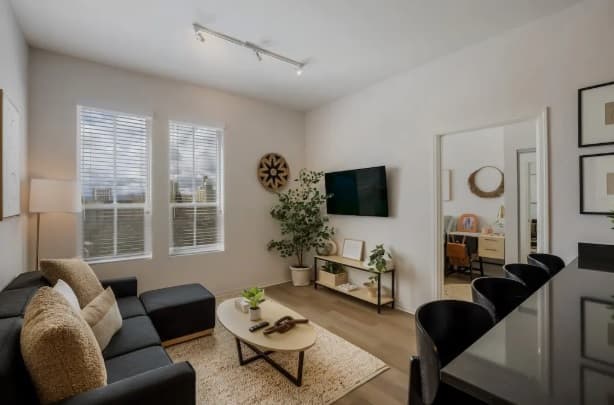 Modern living room by HERE Reno Reno with a black sofa, beige rug, coffee table, TV mounted on the wall, indoor plants, large windows with blinds, and a connected room visible through an open doorway.