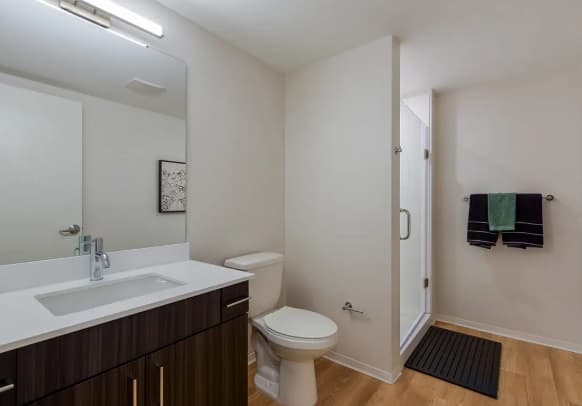 Modern bathroom at HERE Seattle with a large mirror, dark wood vanity, white countertop, toilet, glass shower, towel rack with folded towels, and wood-style flooring. Room is clean and minimalistic.