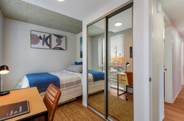 A modern, tidy HERE Seattle bedroom with a single bed, blue and white bedding, a desk with lamp and chair, mirrored closet doors, wall art, and a large window with blinds letting in natural light.
