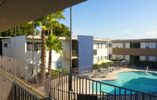 Apartment complex near SDSU with white buildings, outdoor walkways, palm trees, and a swimming pool with lounge chairs under a sunny Huaxtepec blue sky.