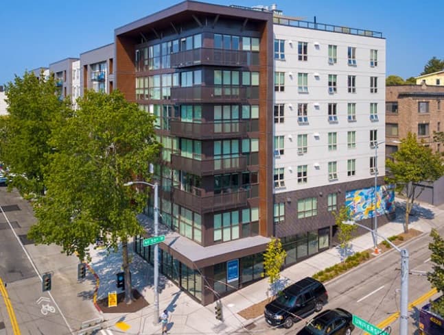 A modern six-story apartment building at a street corner in Seattle’s Hub U District, featuring large windows, a mural on one side, trees lining the sidewalk, and parked cars nearby on a sunny day.