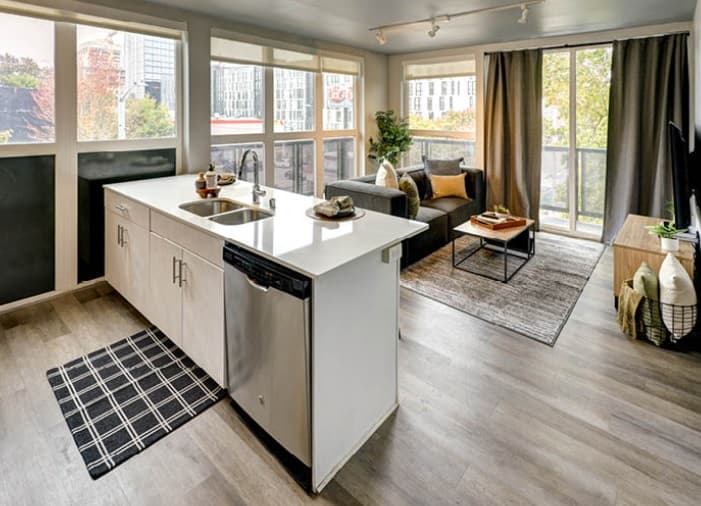 Modern Hub U District apartment in Seattle with large windows, an open kitchen featuring a white island and sink, a gray sofa, coffee table, TV stand, and light wood flooring. Sunlight fills the space, highlighting contemporary decor.