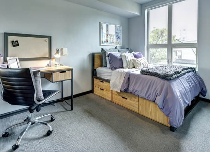A tidy dorm room at Hub U District in Seattle features a neatly made bed with purple and white bedding, storage drawers beneath, a window letting in natural light, a wooden desk with chair, and a corkboard on the wall.