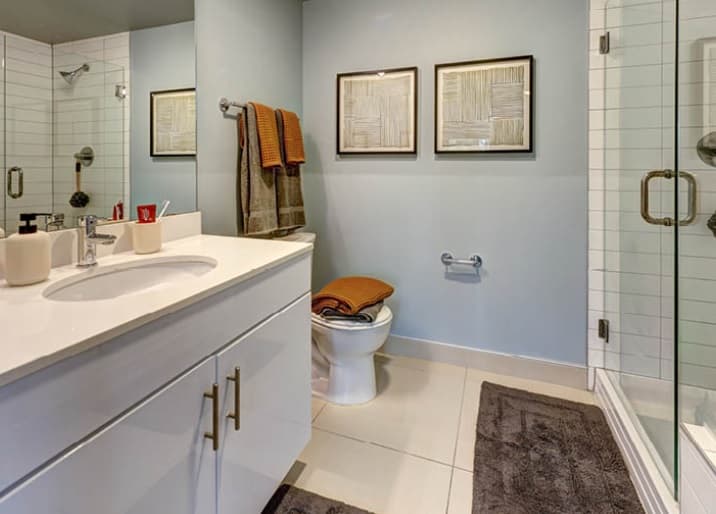 Modern bathroom at Hub U District, Seattle, with a white vanity, double sink and mirror, glass shower on the right, toilet with towels on top, two framed artworks on a light blue wall, and gray rugs on the tiled floor.