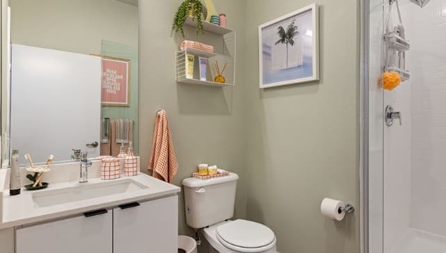 Modern bathroom with a white sink, toilet, and glass shower. Shelves above the toilet hold towels, plants, and decor. Orange towels and accessories add color accents. A framed palm tree photo nods to nearby Hub Los Angeles and the Coliseum.