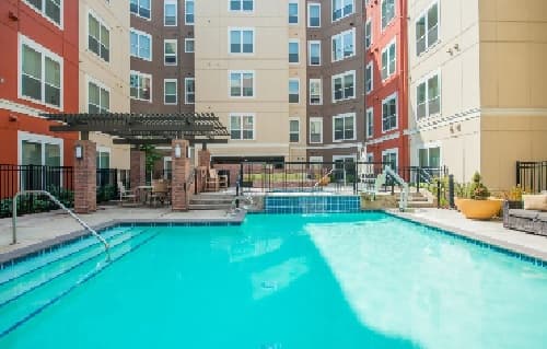 Outdoor swimming pool surrounded by modern Hayward apartments, with poolside seating, tables under a pergola, and potted plants. The bright, inviting area suggests a welcoming community perfect for student accommodation.