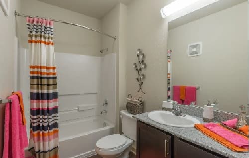 A bathroom in Hayward Student Living features a striped shower curtain, a toilet, a sink with granite countertop, and colorful pink and orange towels. A wall mirror hangs above the sink, with a basket on the toilet—ideal for student accommodation.