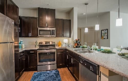 Modern kitchen in Hayward Student Living features dark wood cabinets, stainless steel appliances, granite countertops, a blue rug by the oven, pendant lights, and a breakfast bar set with plates and glasses.
