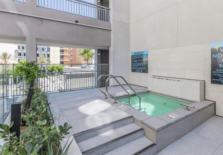 A modern outdoor hot tub with metal handrails, steps leading into the water, and surrounding plants. Safety and warning signs are posted on the beige walls. An urban setting is visible in the background.