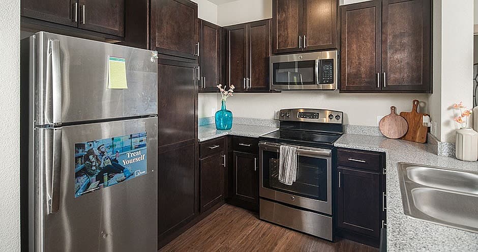 Modern kitchen in Hayward with dark wood cabinets, stainless steel appliances including a refrigerator, oven, and microwave—perfect for student living. Features granite countertops, a double sink, and decorative items like a blue vase and cutting boards.
