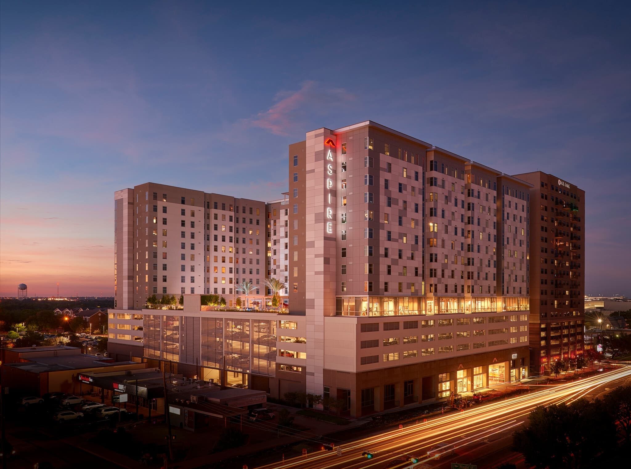 A modern, multi-story apartment building with Aspire in red letters is illuminated at dusk near Ion College Station. The sky is purple and blue, with light trails from cars on the busy street in the foreground.