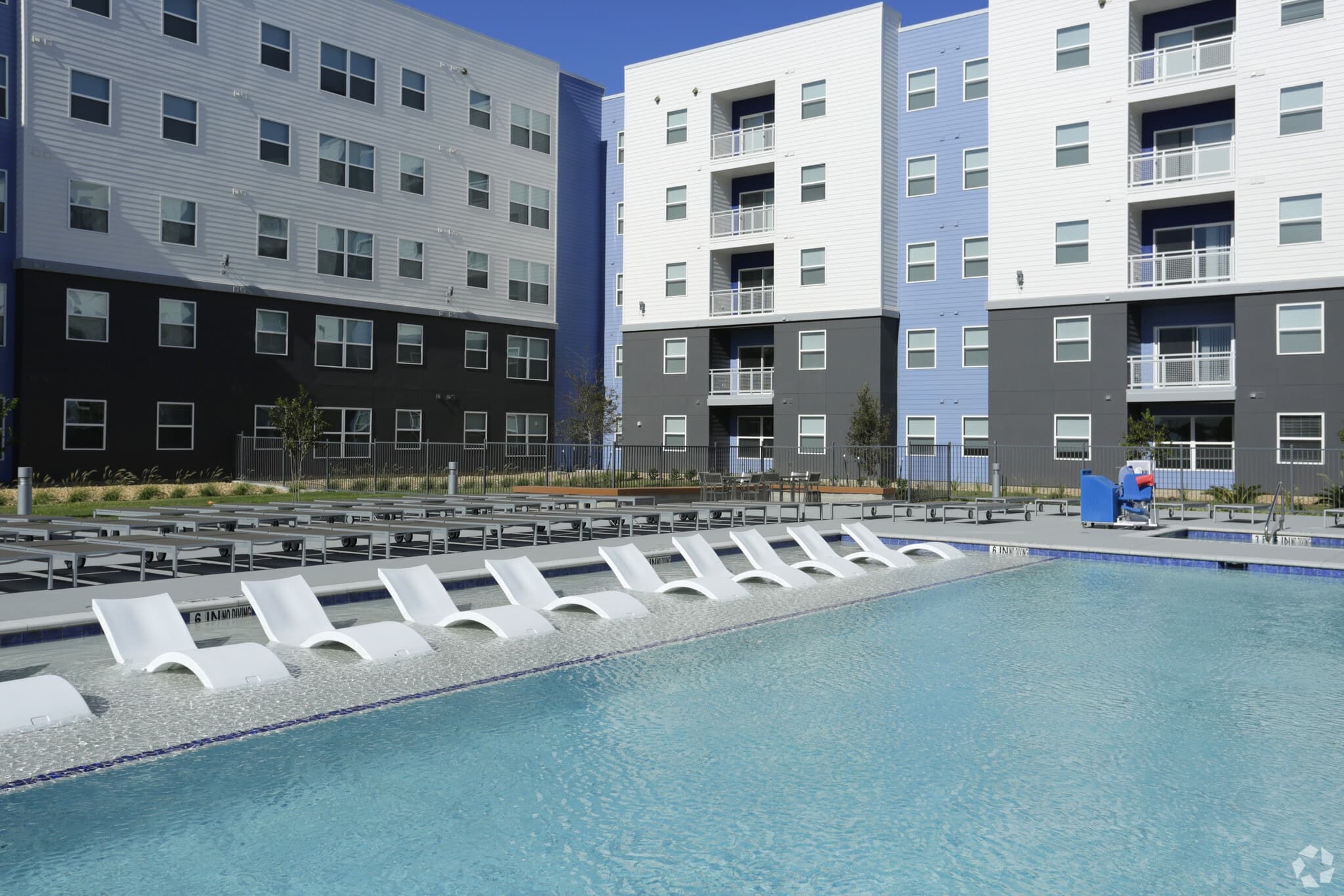 Modern apartment buildings surround a large outdoor swimming pool with white lounge chairs partially submerged in the water. Poolside seating and clear blue sky are visible in the background.