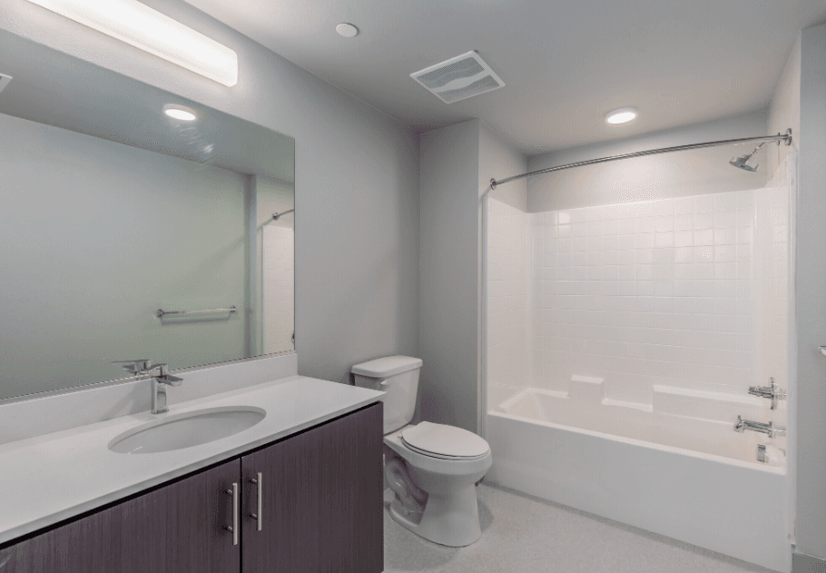 Modern bathroom with a large mirror, a sink with dark cabinets, a toilet, and a white bathtub with a shower and curved curtain rod. The room has light-colored walls and built-in ceiling lighting.