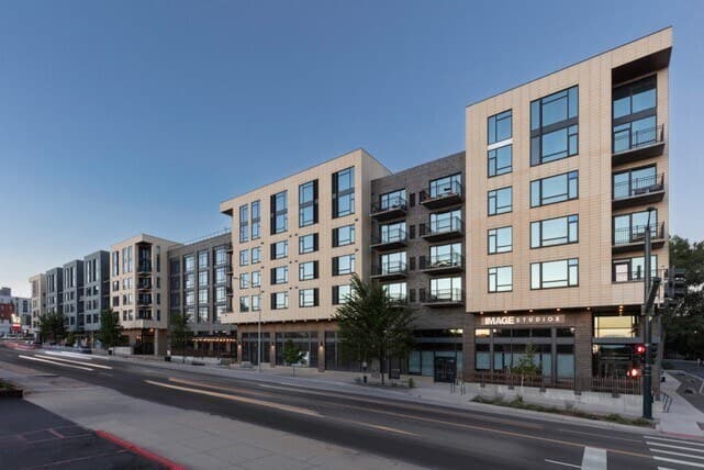 Modern mid-rise apartment buildings with large windows and balconies line a wide city street at dusk. The buildings have retail spaces on the ground floor and trees along the sidewalk.