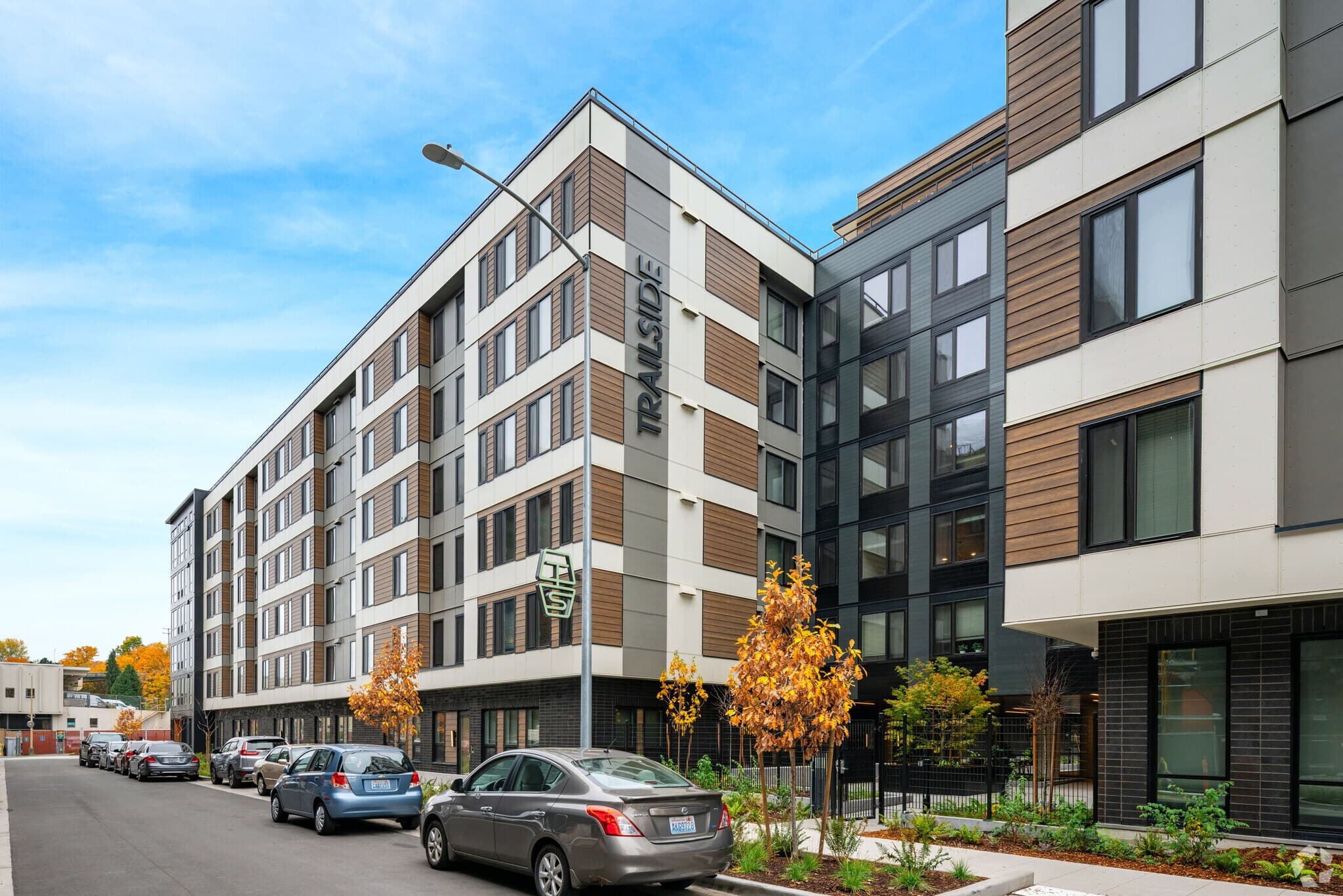 Modern mid-rise apartment building with a sign reading TRAILSIDE, lined with parked cars and small trees along the sidewalk on a clear day.