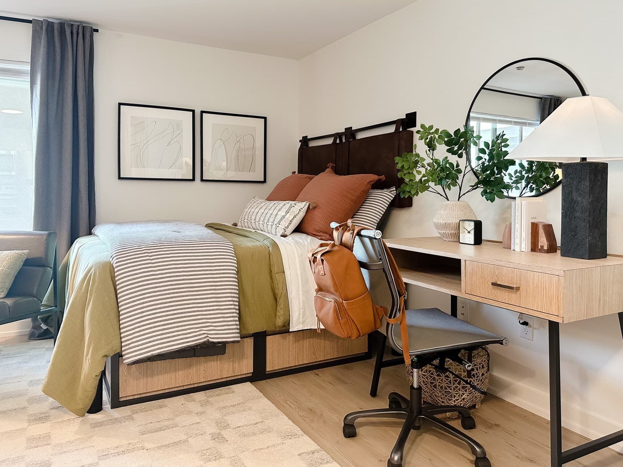 A tidy, modern bedroom in Yugo Seattle student housing features a neatly made bed with striped, earth-toned bedding, a desk with a lamp, books and plants, a chair with a backpack, and minimalistic wall art above the bed.